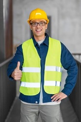 architecture, construction business and building concept - happy smiling male architect in helmet and safety west showing thumbs up gesture at office