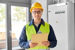 construction business and building concept - happy smiling male electrician or worker in helmet and safety west with papers on clipboard and pencil at electric board