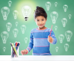 education, school, children, creativity and happy people concept - happy little girl drawing with coloring pencils over green chalk board background and light bulbs