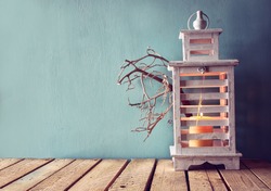 low key image of white wooden vintage lantern with burning candle and tree branches on wooden table. retro filtered image