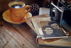 cup of hot coffee next to old photo camera, antique photos and old book on wooden table. vintage filtered image. selective focus