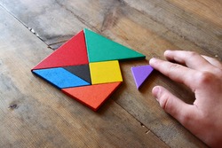 kid's hand holding a missing piece in a square tangram puzzle, over wooden table.
