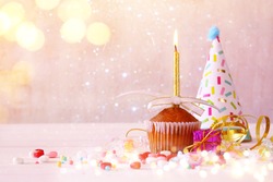 Birthday concept with cupcake and candle, party hat on wooden table. Glitter lights overlay