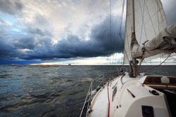 Yacht sailing in an open sea at sunset. Close-up view from the deck to the bow, mast and sails. Dramatic stormy sky, dark clouds, winter cyclone, rough weather. Epic seascape. North sea, Norway
