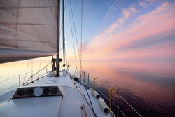 White yacht sailing in an open sea at sunset. Close-up view from the deck to the bow. Clear blue sky with glowing pink clouds reflecting in a still water. Idyllic seascape. Cruise, travel destinations
