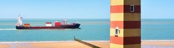 Cargo container ship sailing in the sea. Coast of Vlissingen, the Netherlands. Dunes and a striped lighthouse. Freight transportation, nautical vessel, logistics, industry, environment. Panoramic view