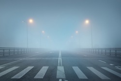 An empty illuminated asphalt road (highway) in a thick fog. Pedestrian walkway, crossing, street lights. Dangerous driving, walking, cycling, traffic laws concepts