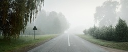 An empty highway (asphalt road) through the fields and forest in a thick fog at sunrise. Atmospheric landscape. Idyllic rural scene. Darkness, fall season, fickle weather, dangerous driving, road trip