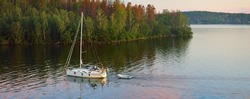 Sloop rigged yacht with inflatable boat sailing at sunset. Panoramic aerial view of the forest lakeshore. Moonrise, glowing clouds. Mälaren lake, Sweden. Summer vacations, nature, sailing, cruise