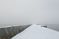 Panoramic view of Baltic sea from sandy shore. Promenade to the lighthouse, breakwaters. Thick white fog, mist, snow. Waves, water splashes. Seascape. Monochrome winter scenery