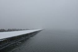 Panoramic view of Baltic sea from sandy shore. Promenade to the lighthouse, breakwaters. Thick white fog, mist, snow. Waves, water splashes. Seascape. Monochrome winter scenery