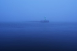 Panoramic view of Baltic sea. Port entrance, lighthouse, breakwaters. Thick fog, mist, blue light. Waves, water splashes, long exposure. Seascape. Monochrome scenery. Danger, safety concepts