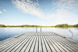 Wooden pier near the river (lake). Evergreen trees in the background. Clear blue sky, reflections on the water. Idyllic summer landscape. Ecotourism, recreation, ecological resort, spa, sauna. Finland