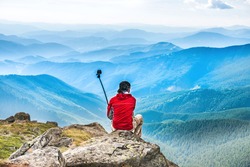 Young man on the top of mountain taking picture with camera on selfie stick