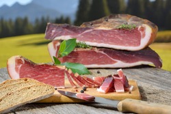 Typical South Tyrolean bacon snack with local rye bread lying on a rustic table in front alpine pastures and mountains of the alps