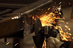 A metal worker grinds away welded  metal spatter and cleans the surface of a welded surface, causing a spray of sparks.