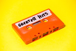 A vintage cassette tape (obsolete music technology), orange on a yellow surface, angled shot, carrying a label with the handwritten text Greatest Hits.
