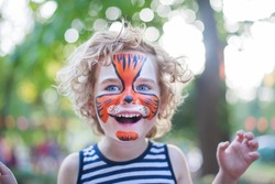 smiling boy with curly hair and face art painting like tiger, little boy making face painting, halloween party, child with funny face painting, little cute boy with faceart on birthday party close up