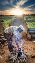 African man lifting the lid from an old burned tea pot standing on the fireplace in an African village