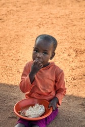 Small African child eating on the ground a plate of meat and sorghum