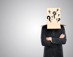 Businessman with folded arms and cardboard box with question marks covering head on grey background. Confusion concept