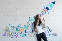 Attractive european woman celebrating success on brick wall background with creative drawn space ship. Successful young entrepreneur