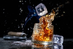 Icecube splashing to glass of whiskey. Copy space, black background. Smoking cigar and whiskey, brandy, cognac, alcoholic drink .