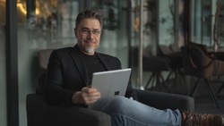 Business portrait - businessman using tablet computer in office lobby or on cafe terrace. Happy middle aged man, entrepreneur working online.