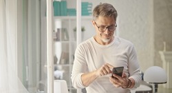Happy older good looking casual man using phone at home, smiling. Portrait of mid adult, mature age man in glasses,