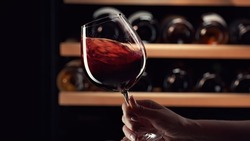 Close up female hand swirling red wine in wine glass. Wine expert tasting, rating and drinking wine, bottles in background.