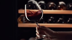 Close up female hand swirling red wine in wine glass. Wine expert tasting, rating and drinking wine, bottles in background.