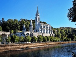 A picturesque summer view of the Basilica of Lourdes and the Gave de Pau under a clear blue sky.