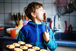 Cute little happy blond school kid boy baking carrot blueberry muffins in domestic kitchen, indoors. Funny lovely healthy child having fun, tasting sweet dough. Little helper
