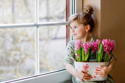 Little toddler girl sitting by window with blossoming pink hyacinth flowers. Happy child, indoors. Mother's day, valentine's day or birthday and spring concept.