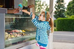 Cute little toddler girl choosing and buying ice cream in outdoor stand cafe. Happy preschool child with glasses looking at different sorts of icecream. Sweet summer dessert.