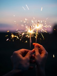 Male and female hands holding sparkling fireworks to the sky at twilight against sunset sky