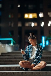 Woman with a smartphone sits on long stone steps in the night city