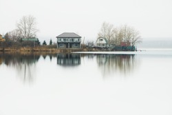 Houses and trees and their reflections in the calm water of lake. Misty morning in early spring. South Ural, Russia