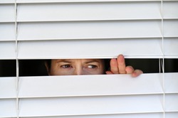 Suspicious person looking outside home window from behind Venetian curtains. Concept photo of person that have Pistanthrophobia and Agoraphobia, a person who don't trust fear of leaving the house. 