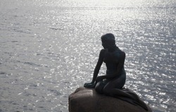 Dark silhouette of statue of little mermaid on stone against water waves 