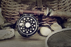 Vintage concept, to include grain effect, of a wet antique fly fishing reel, rod, landing net, artificial flies and rocks in front of creel with rustic wood underneath. Layout in horizontal format. 
