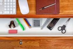 High angle shot of open desk drawer with basic work items inside. Cherry wood desktop has computer keyboard, mouse and executive notepad with pen. Wooden oak floors underneath desk. 