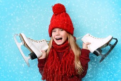 Cheerful little girl in warm sweater and hat holding figure skates. Blue background. 