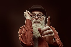 A wise old man with a long white beard in a bowler hat and glasses gives advice and shows his index finger. Black background. 