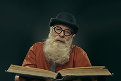 An intelligent old man in glasses and bowler with a long gray beard holds an old big book and looks at the camera. Old age wisdom.