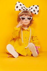 Lovely little child girl in fashionable dress, big bow and sunglasses poses on a bright yellow background. Kid's fashion. 