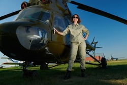Full length portrait of a professional female commercial aviation pilot woman in uniform and sunglasses poses happily smiling in the background of a helicopter on the airfield. Aviation.