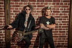 Two punk rock musicians in concert costumes posing with an electric guitar near a brick wall. Youth alternative culture. Grunge style. 