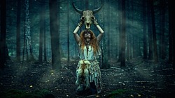 Female shaman in an ethnic dress doing a mysterious ritual holding an animal skull in her hands. Dark gloomy forest background. Black magic concept, fantasy. Paganism. Halloween.