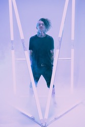 Cool punk rock star with dreadlocks in black clothes poses among neon lamps with his hands in his pockets surrounded by haze. Space punk rock music. Youth alternative culture. Full length portrait.